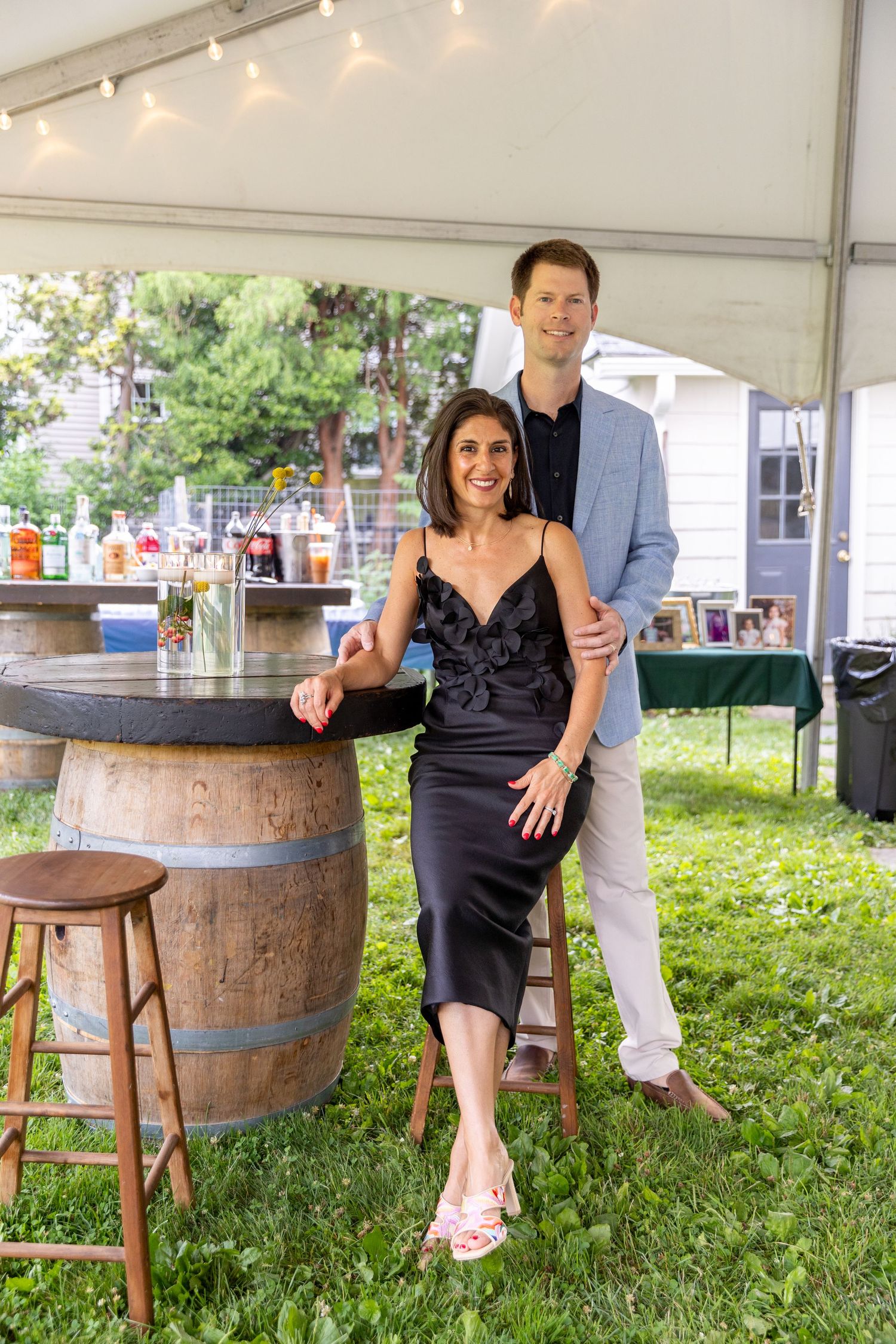 Couple posing together at an outdoor party under a tent near a wooden barrel bar with string lights.