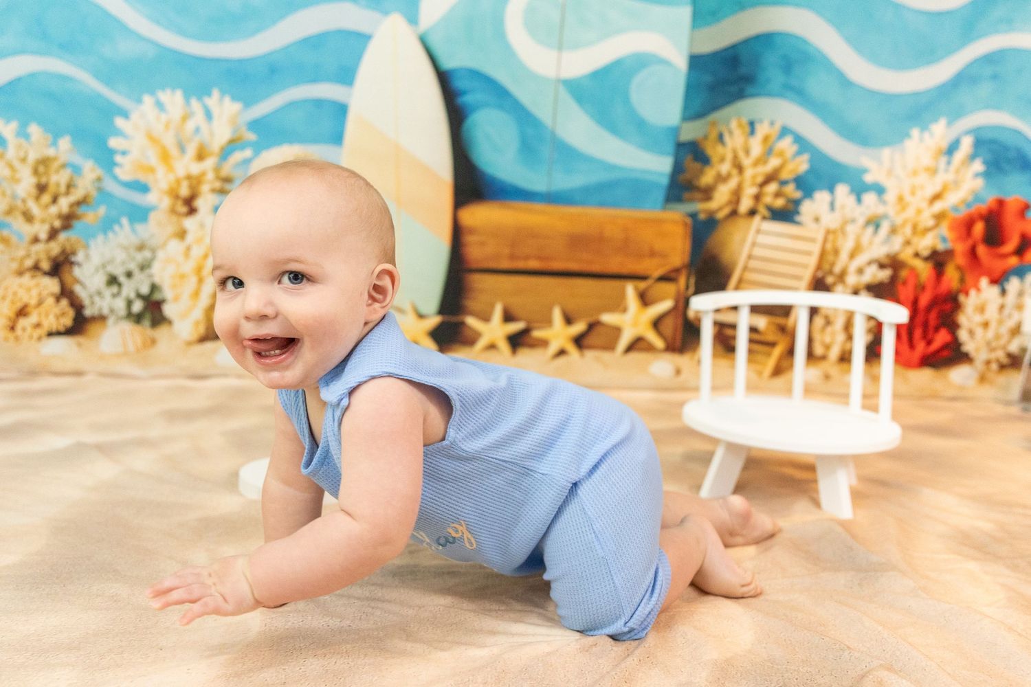 Happy baby boy in blue romper crawling on sand in front of beach-themed birthday backdrop.