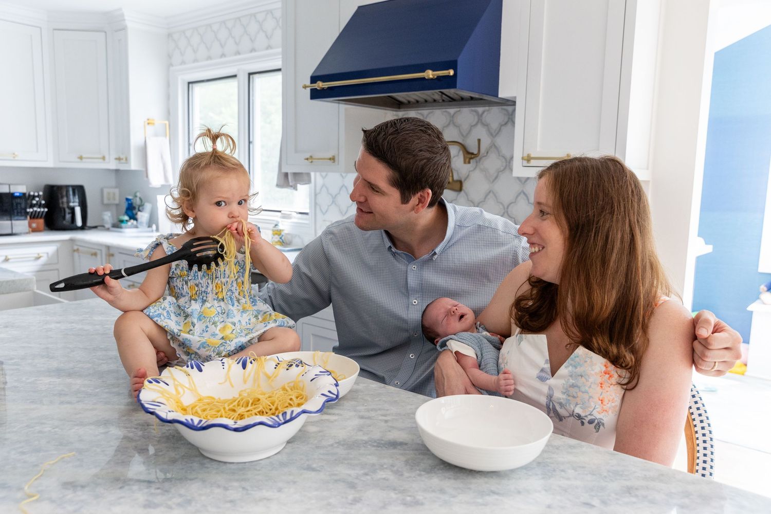 Young family with toddler and newborn enjoying pasta together in a bright white kitchen.