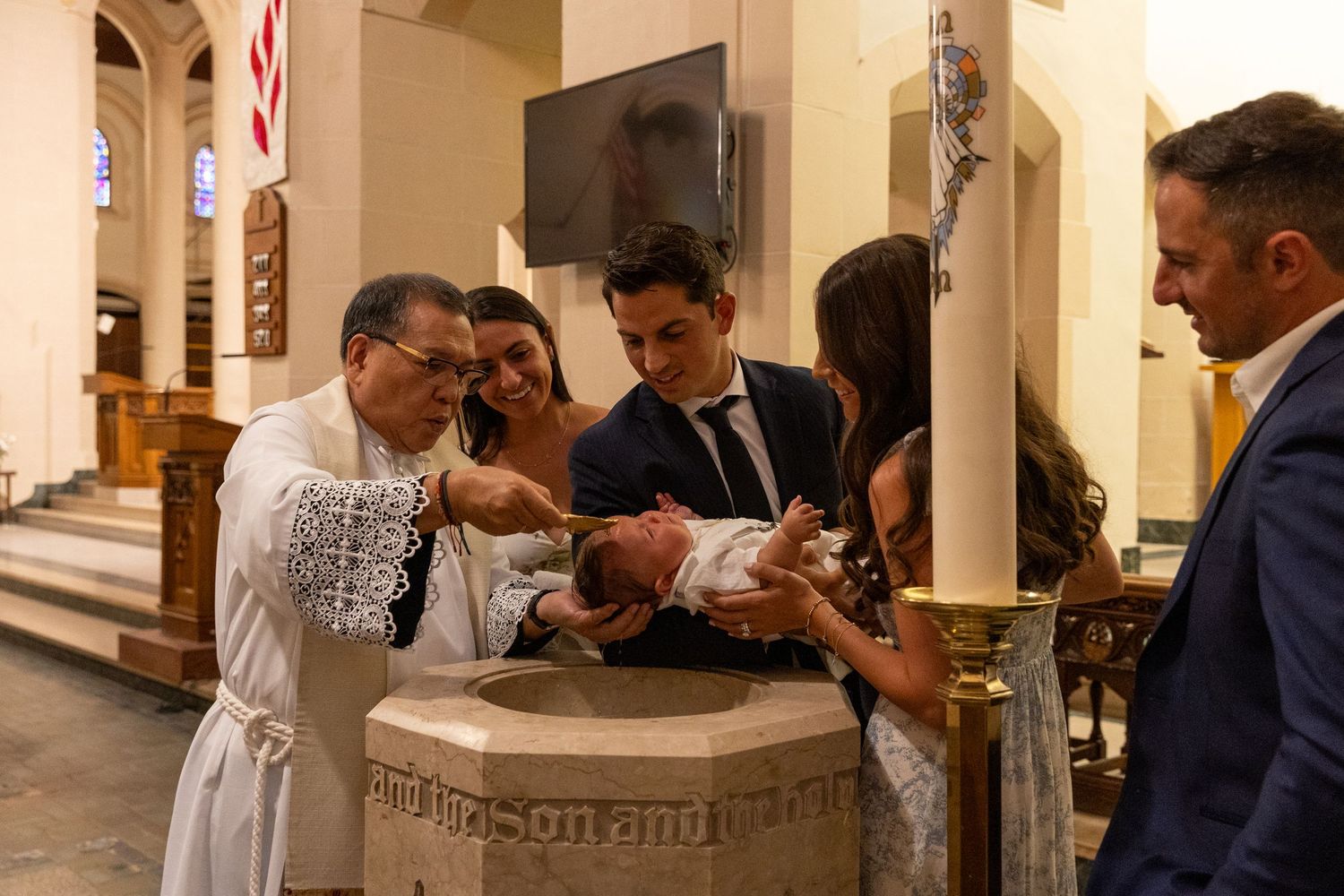 A priest performs a Catholic baptism ceremony at a stone font surrounded by family members inside a church.