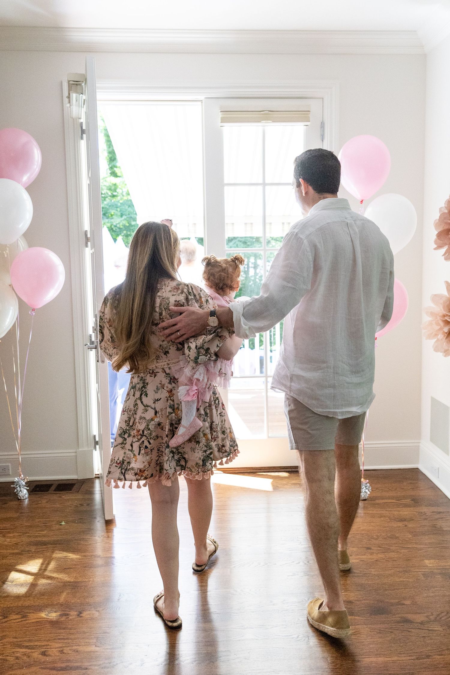 Parents carrying their toddler daughter toward the door surrounded by pink and white birthday balloons.