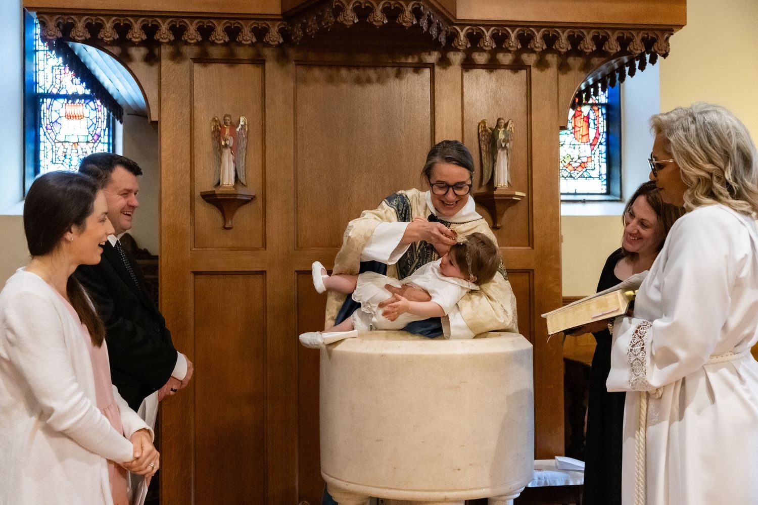 A clergy member baptizes an infant at a white baptismal font while smiling guests look on in a church.