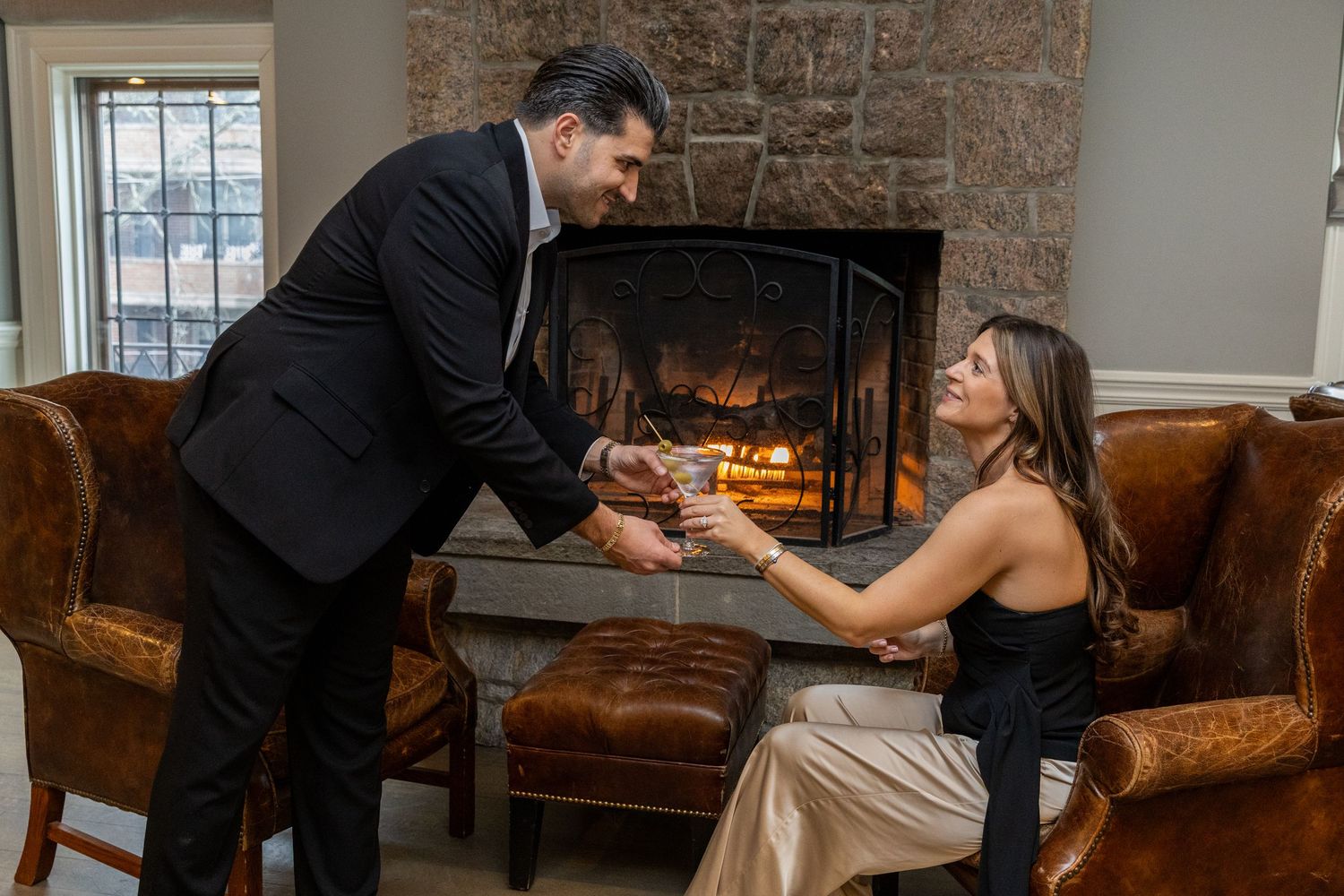 A man in a black suit offers a drink to a woman seated by a stone fireplace in a cozy room.