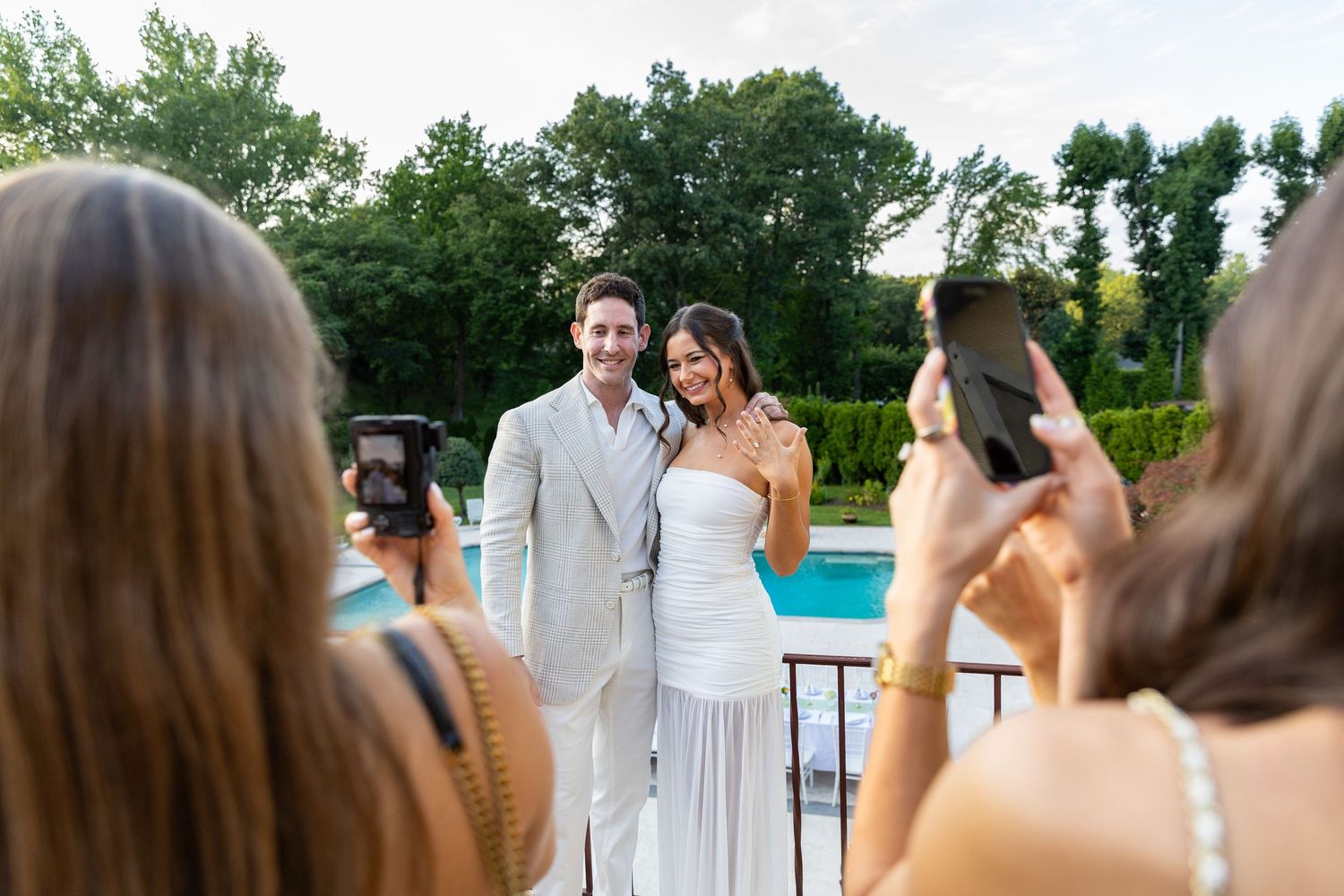 Engaged couple posing poolside while guests photograph them with smartphones.