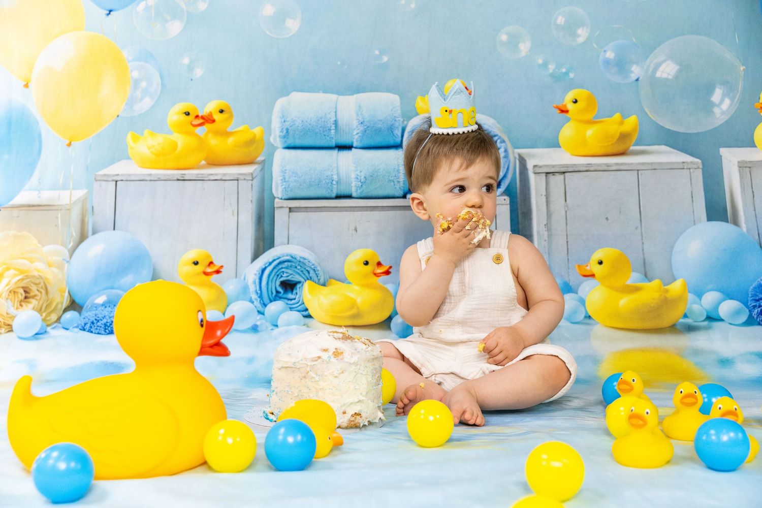 One-year-old baby eating birthday cake surrounded by rubber ducks and colorful balloons.