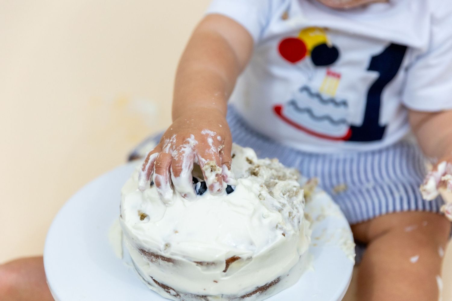 Close-up of baby hands covered in white frosting smashing first birthday cake in blue striped outfit.