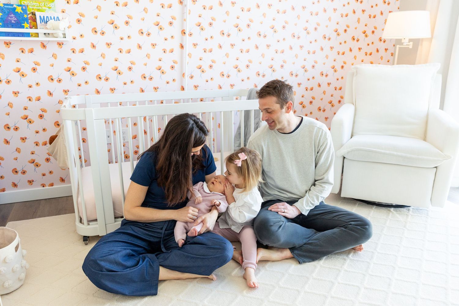 Young family sitting on nursery floor with newborn and toddler near orange floral wallpaper.