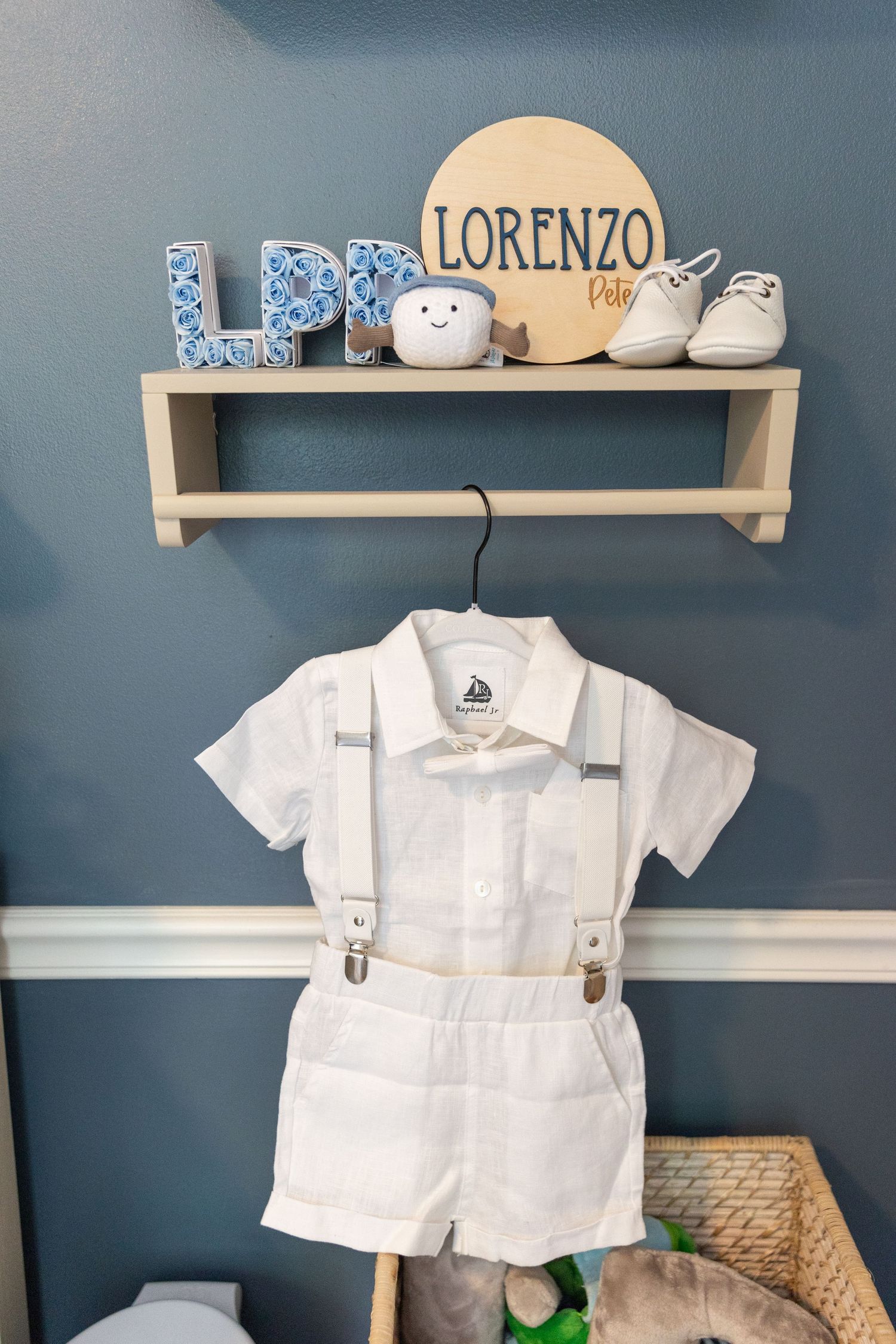 Baby nursery shelf with Lorenzo name sign above white suspender outfit on hanger.