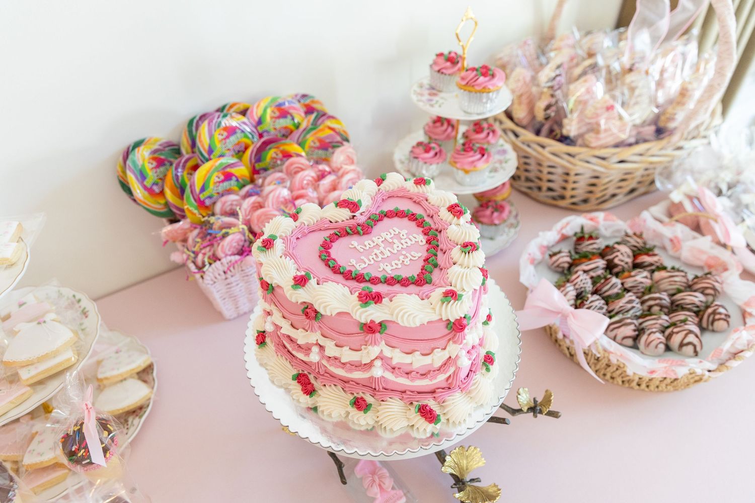 Elegant pink heart-shaped birthday cake surrounded by lollipops and chocolate treats on a dessert table.