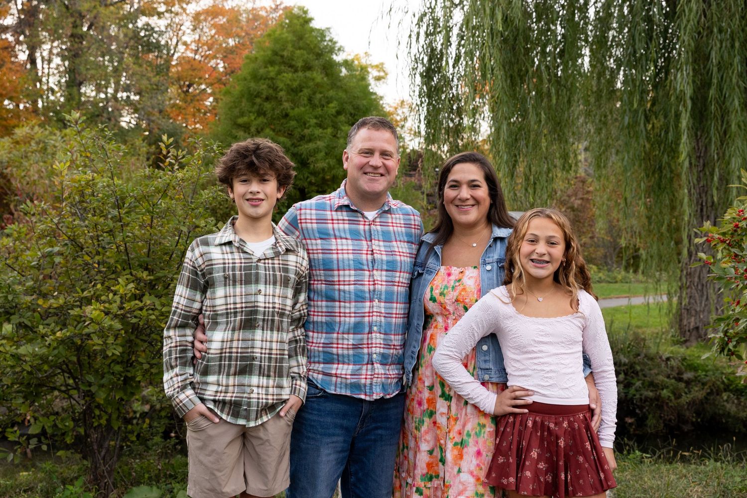 Family of four smiling together outdoors with a weeping willow and fall trees behind.
