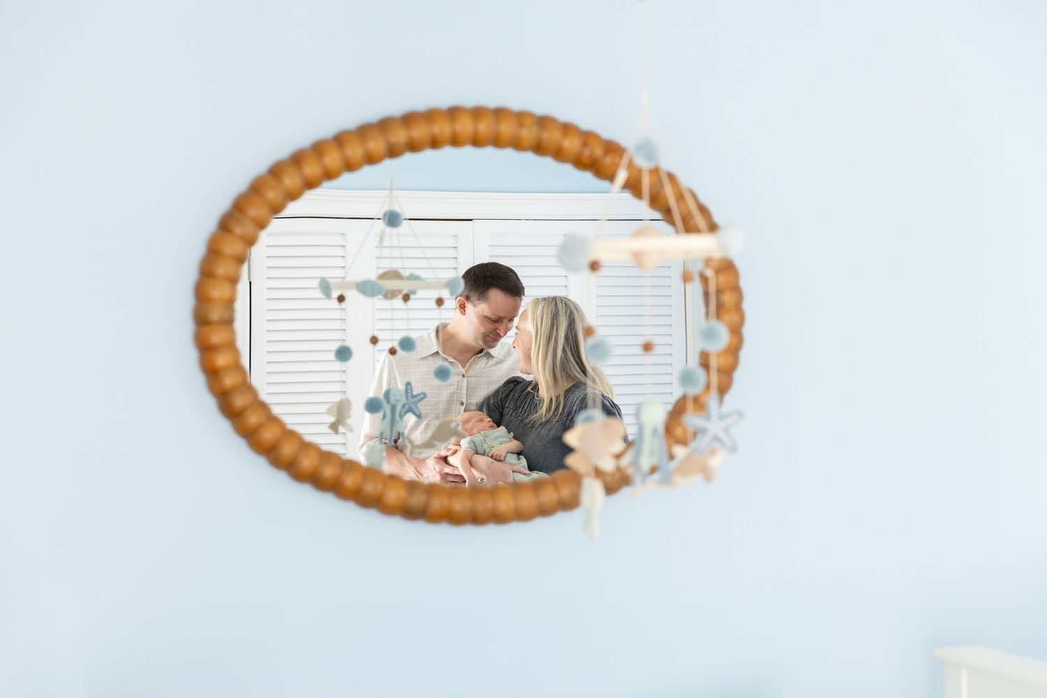 A couple shares a tender kiss reflected in a round rattan mirror decorated with a baby mobile.