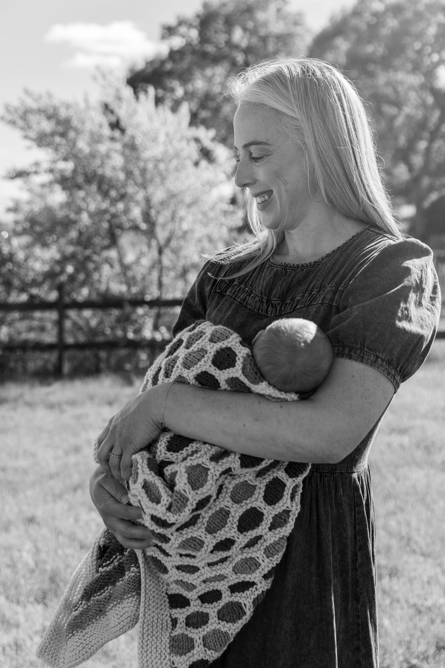 Smiling blonde mother in black dress cradling wrapped newborn baby outdoors in black and white photo.