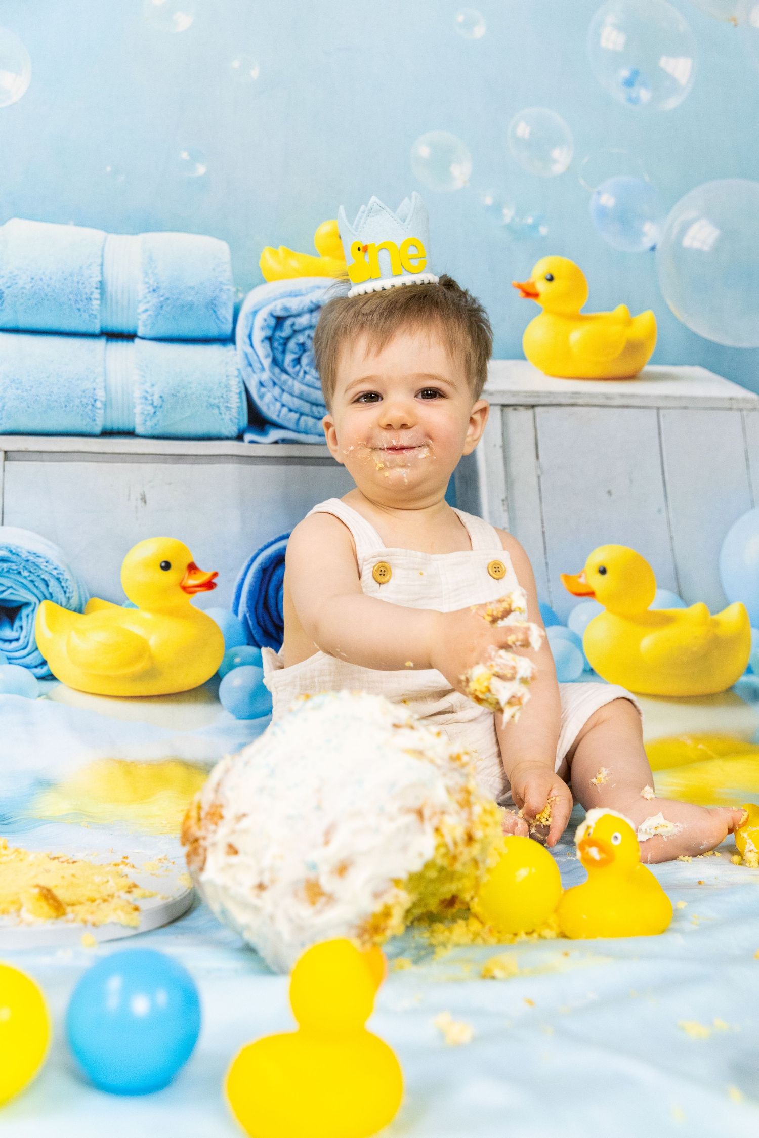 Happy baby covered in cake during rubber duck themed first birthday cake smash session.