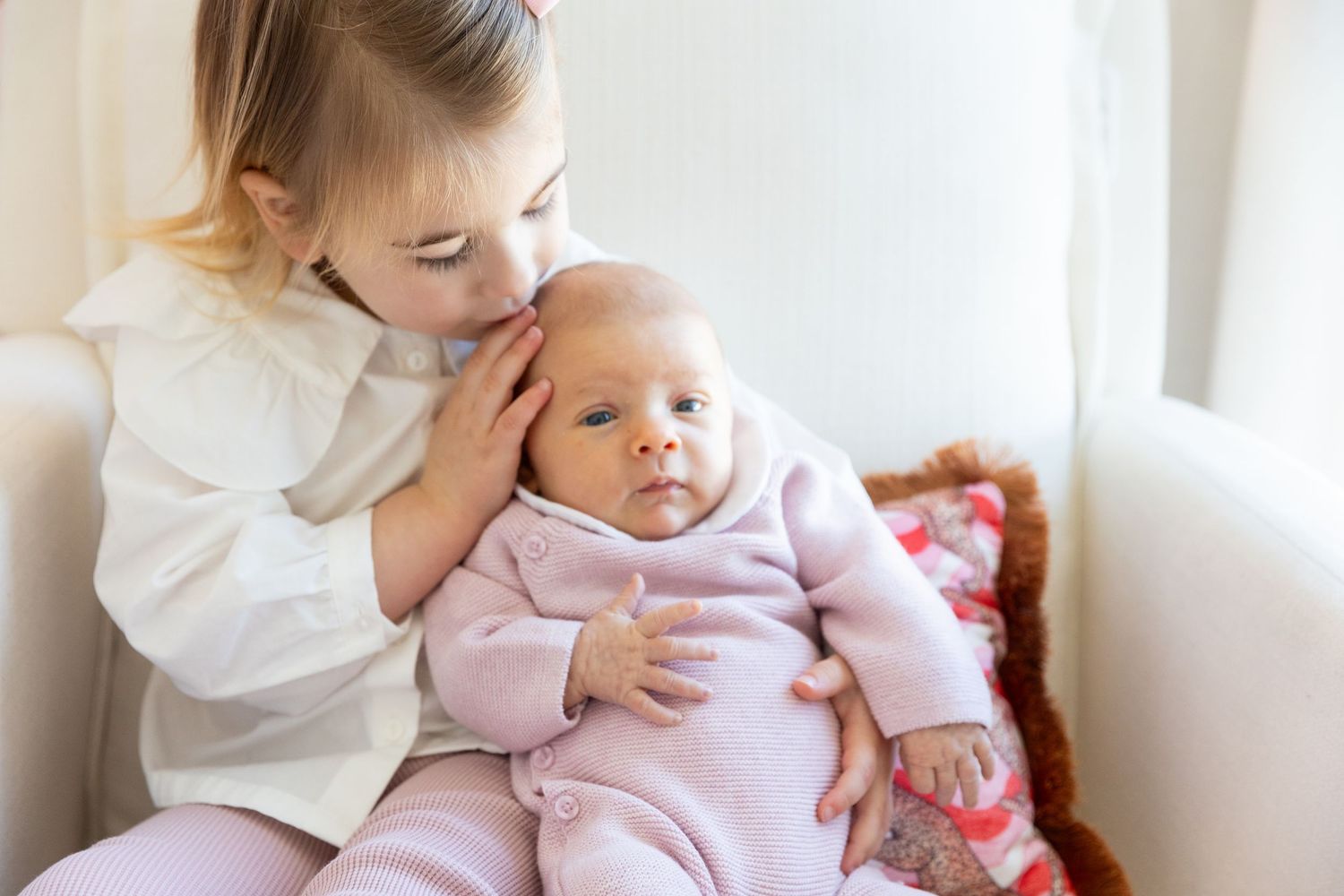 Gentle toddler girl in white dress carefully holding a newborn baby in pink outfit.