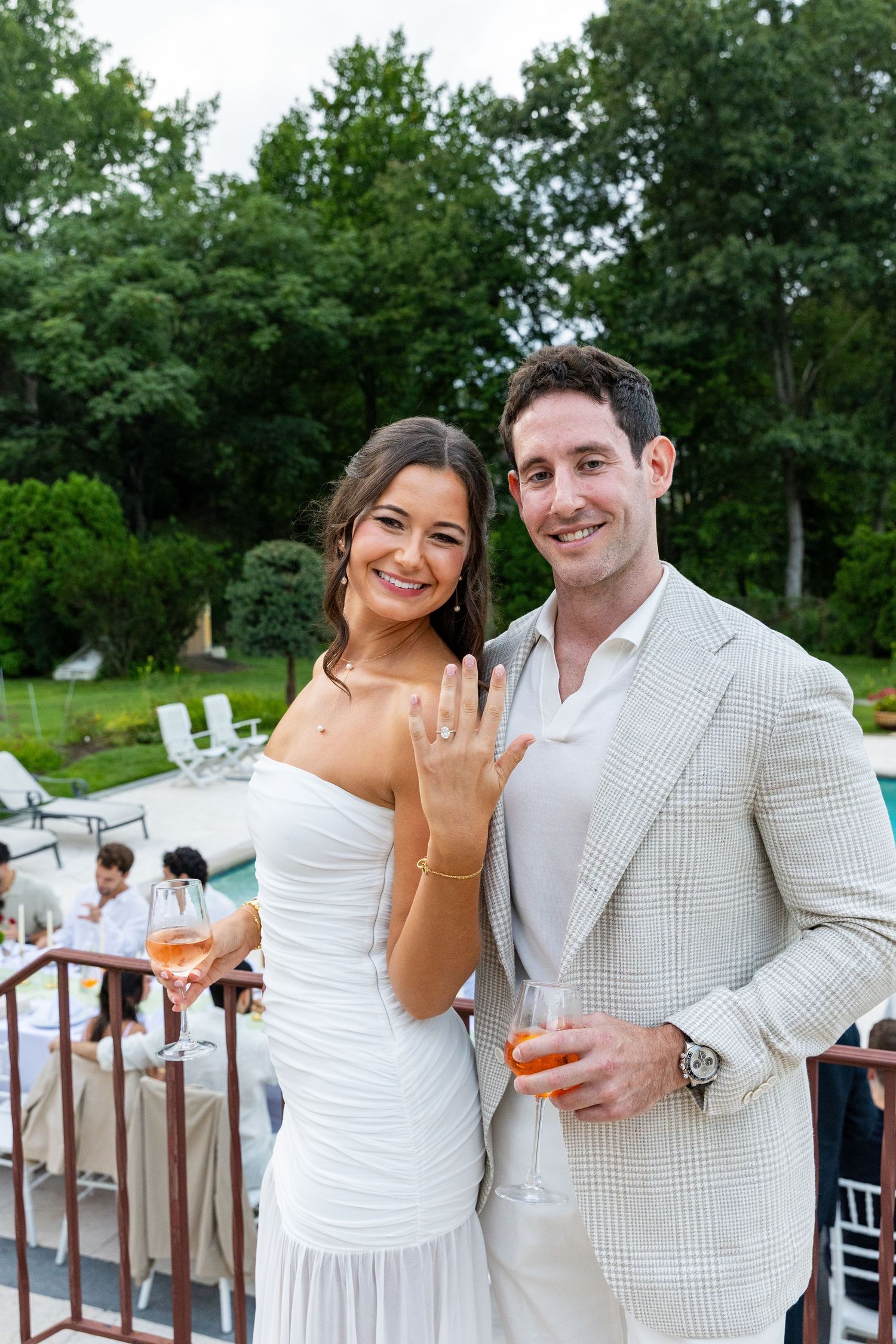 Smiling bride to be showing engagement ring beside groom in tan suit near pool.