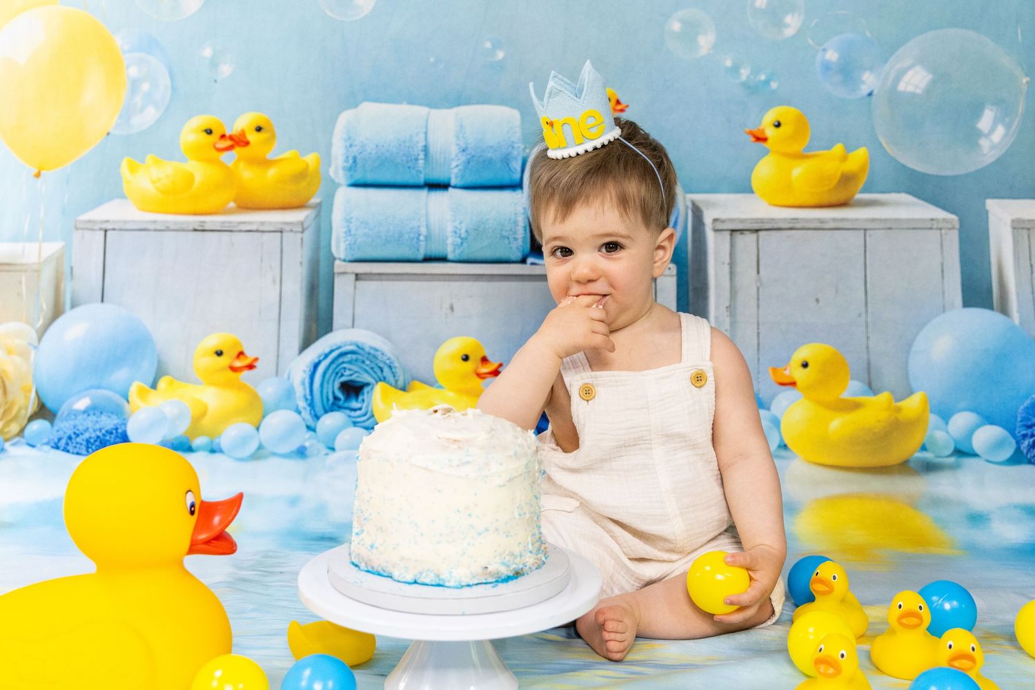 Baby wearing crown sits with white birthday cake in rubber duck themed photo session.