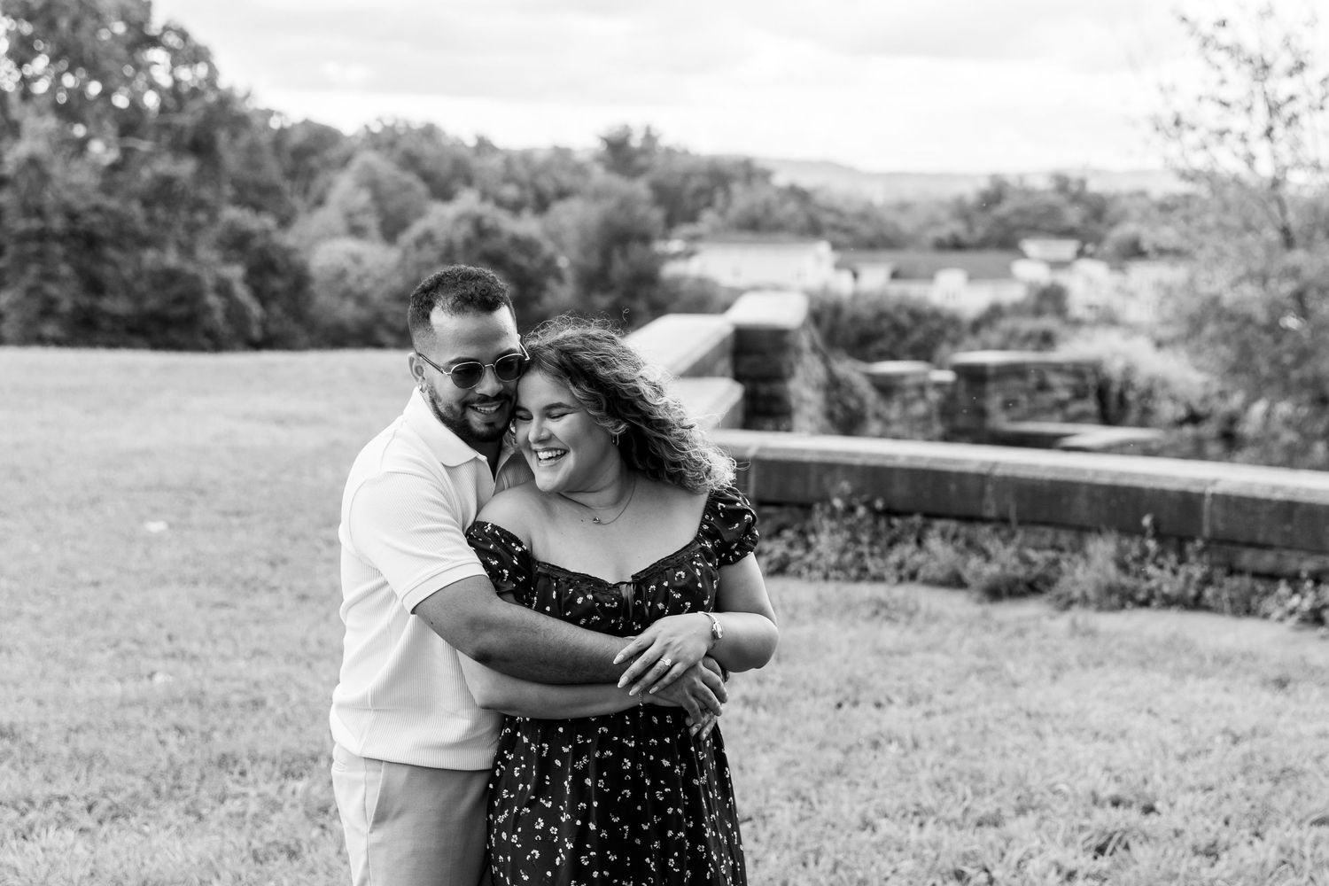 Black and white photo of smiling couple embracing outdoors with scenic countryside background.
