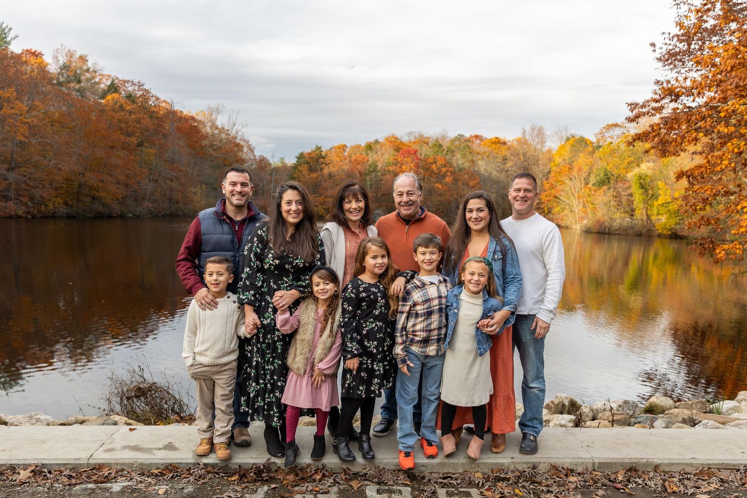 Large extended family group portrait by a reflective lake with vibrant fall foliage.