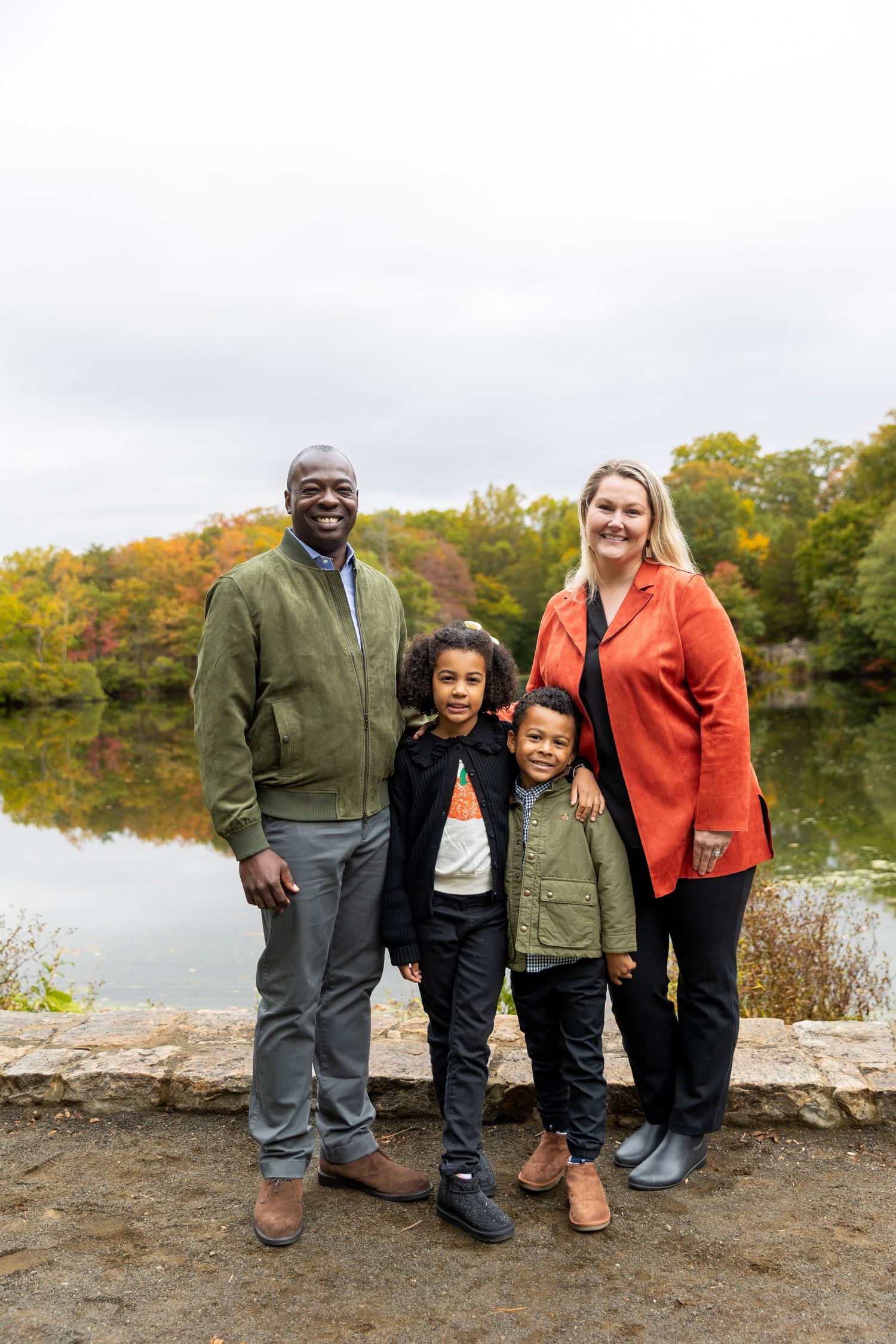Interracial family of four smiling together by autumn lake with colorful fall foliage background.