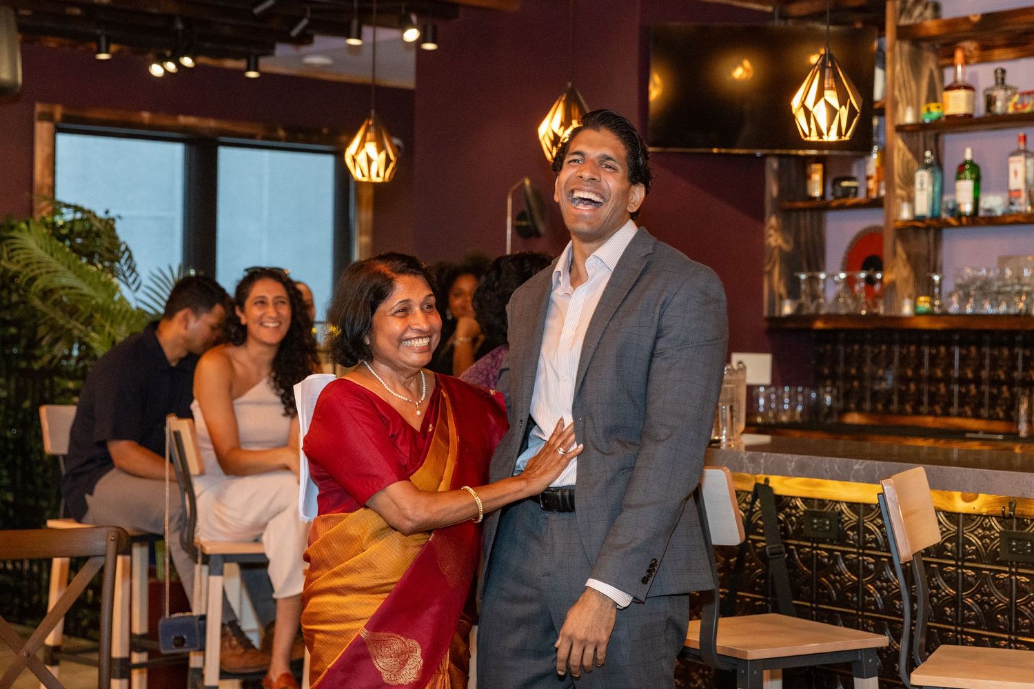 Woman in red saree and man in grey suit laughing together at an indoor birthday party celebration.