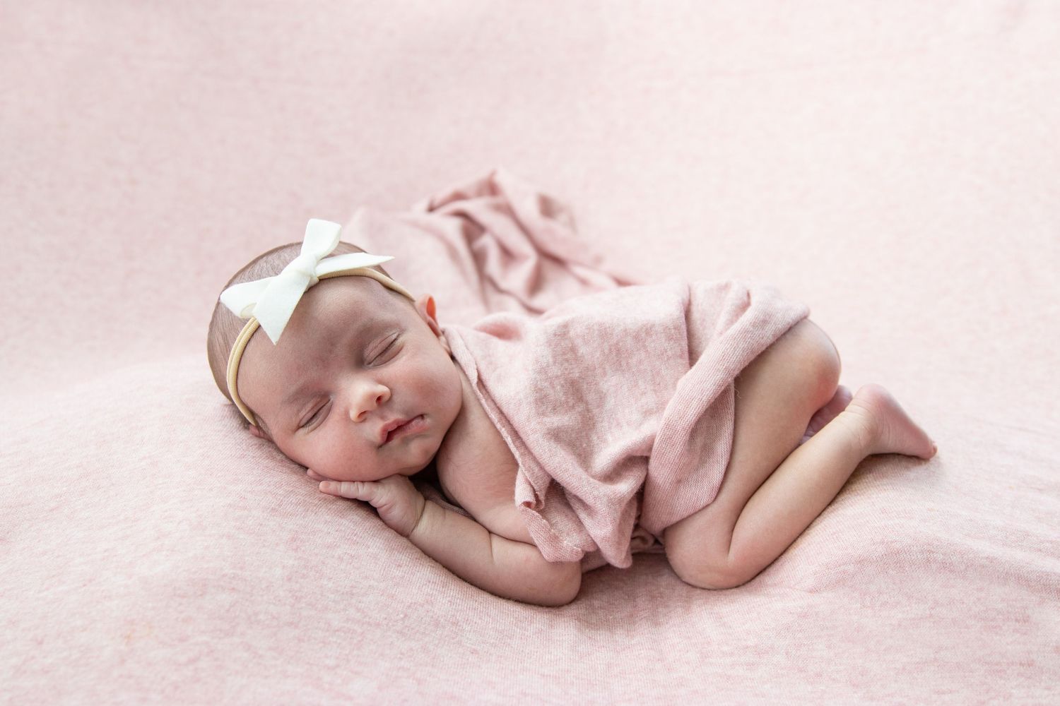 Peaceful newborn baby girl sleeping wrapped in pink blanket with white bow headband.