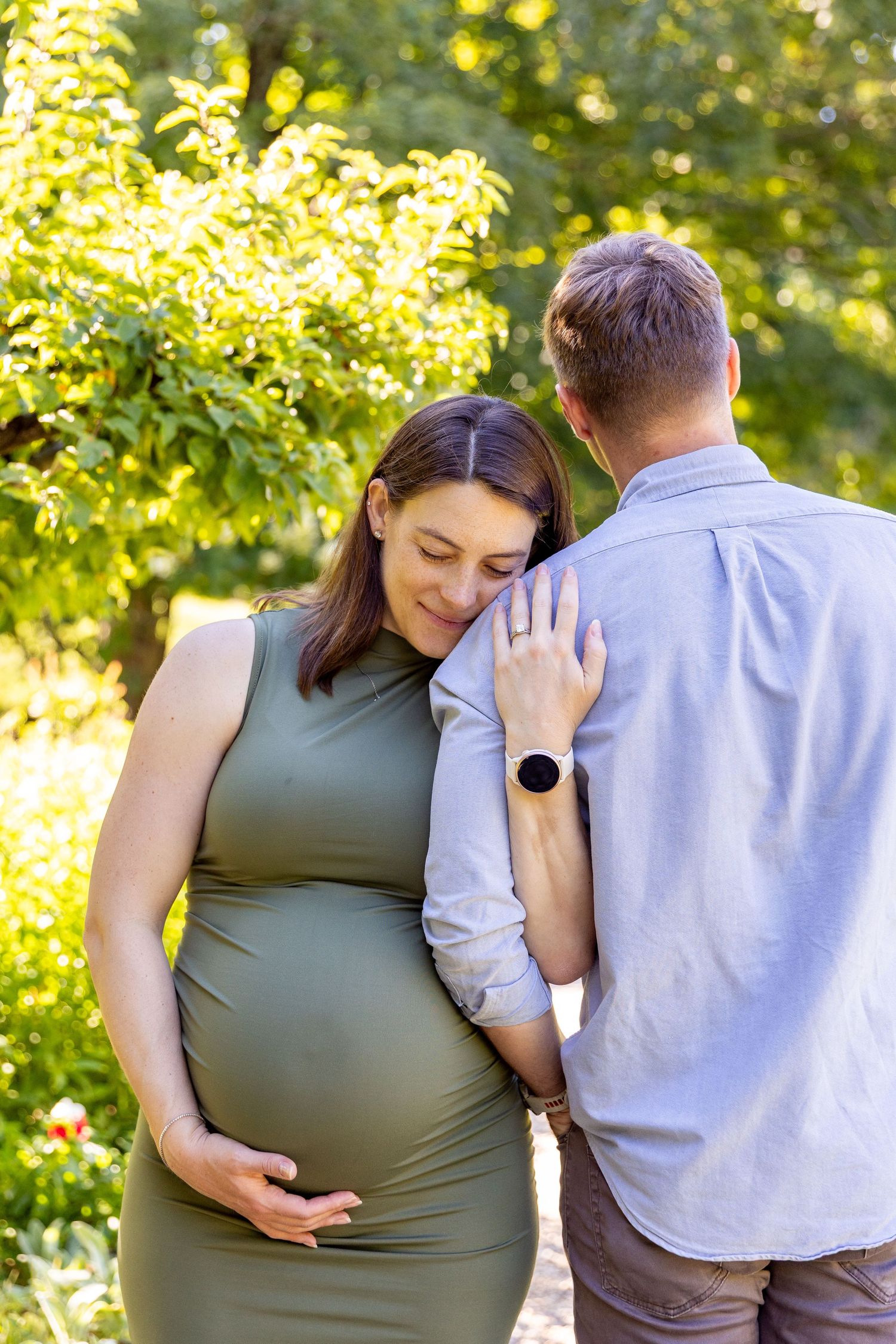 Pregnant woman in olive dress leans on partner outdoors in sunny green garden during maternity photo session.