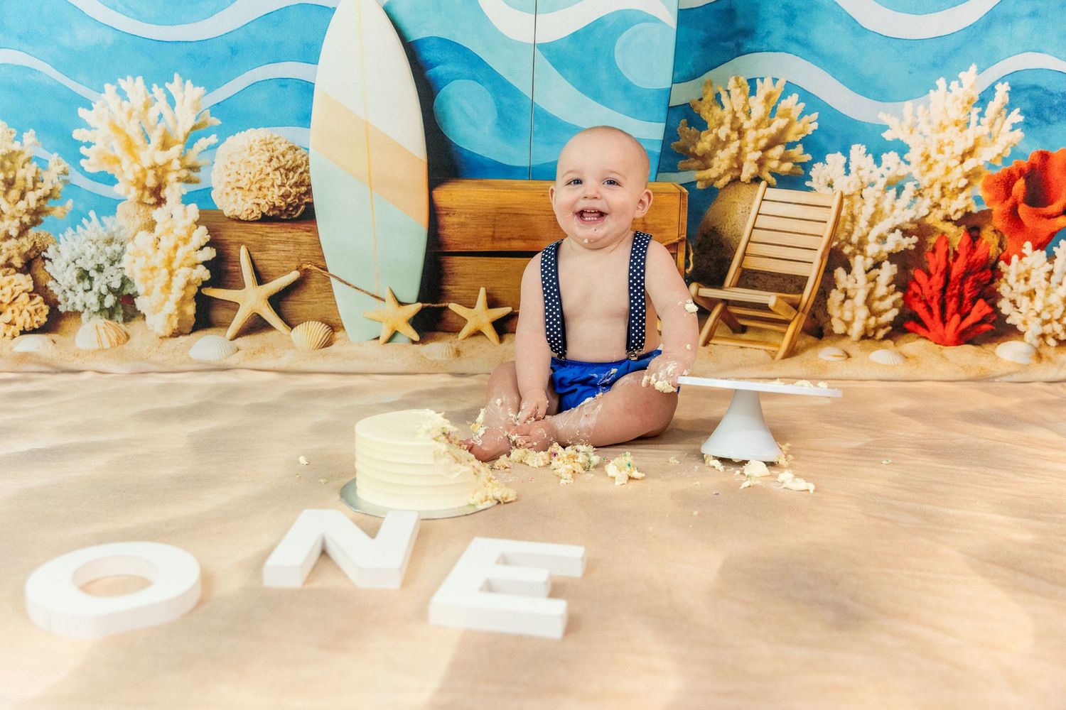 Smiling baby boy in suspenders doing a cake smash on sand with beach-themed ONE birthday decor.