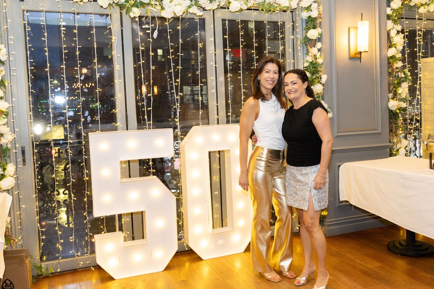 Two women posing together in front of a large illuminated 50 sign at a milestone birthday celebration.