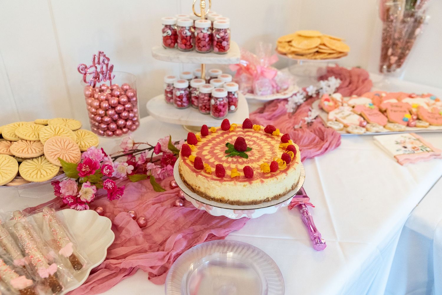 Elegant baby shower dessert table with cheesecake, pink candies, cookies, and floral decorations.