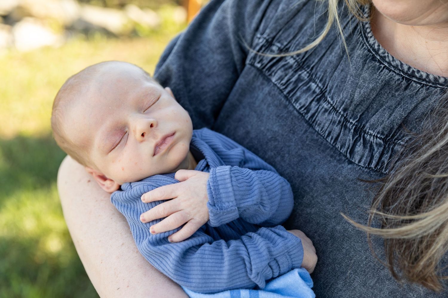 Sleeping newborn baby in a blue outfit cradled gently in a mothers arms outdoors.