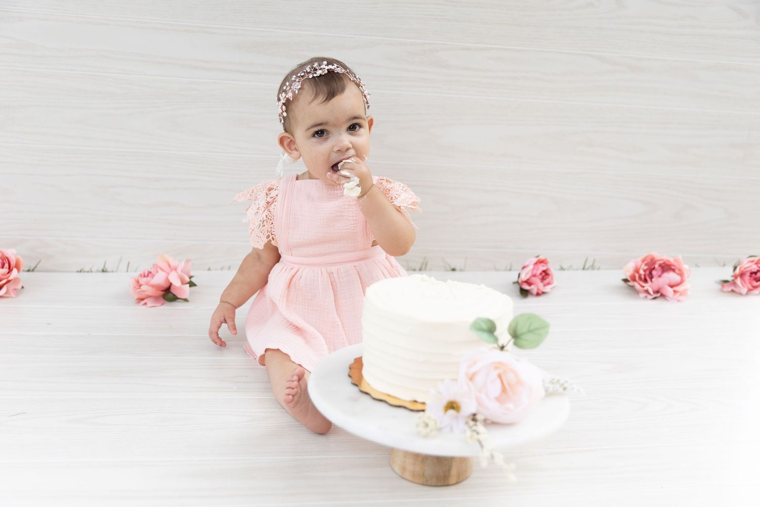 Baby girl in pink dress eating first birthday white cake surrounded by pink roses.