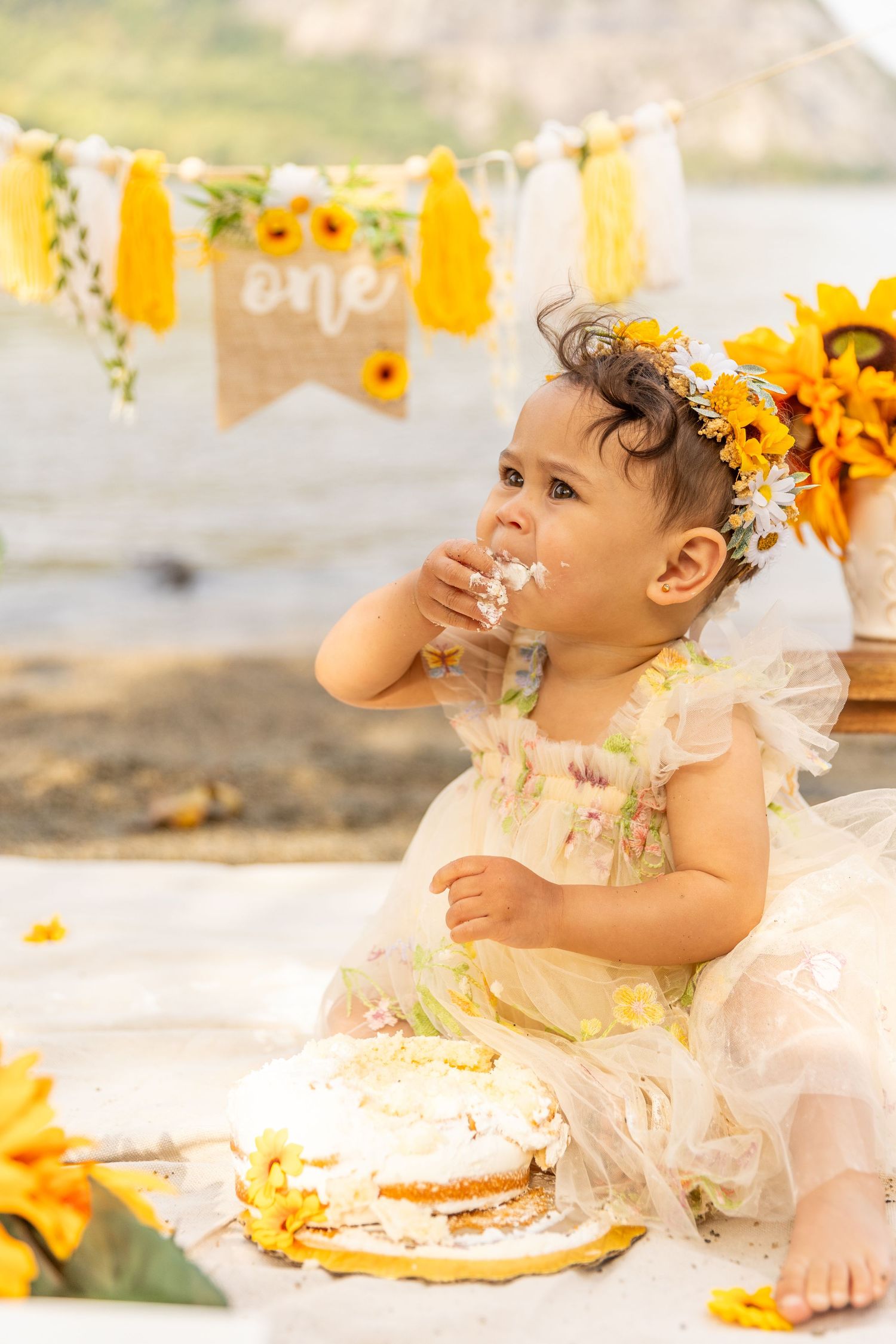 Baby girl in floral crown and white dress smashing cake at sunflower-themed first birthday party.