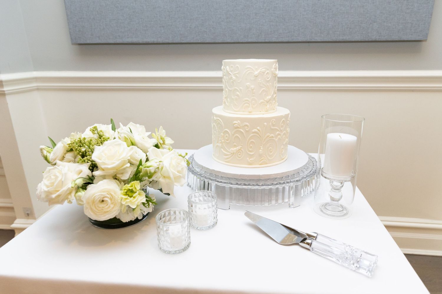 An elegant two-tier white wedding cake with lace detailing displayed beside white roses and candles.