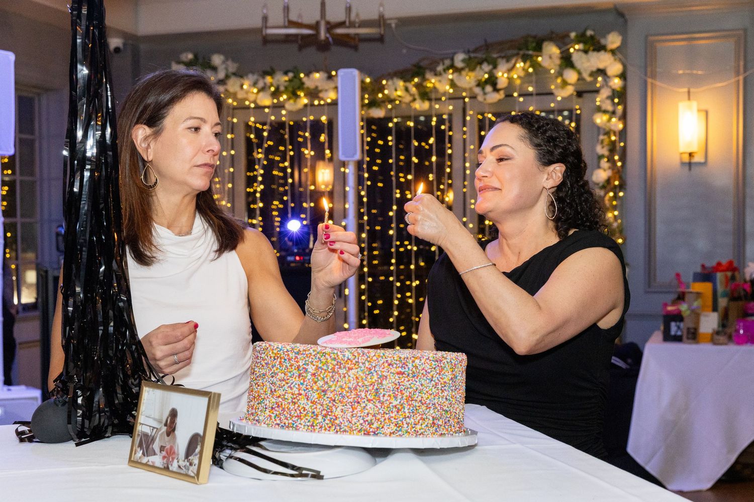 Two women lighting candles on a colorful sprinkle birthday cake at an elegant party celebration.