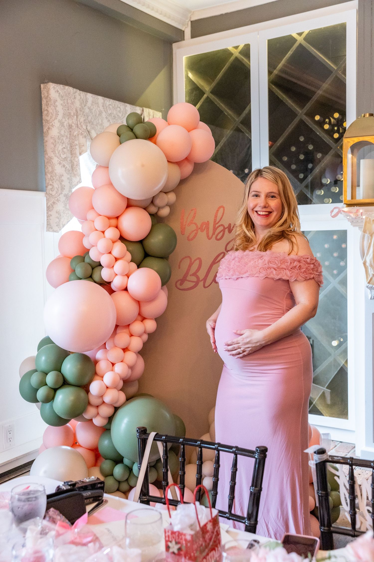 Pregnant woman in pink gown smiling at baby shower with pink and green balloon garland backdrop.