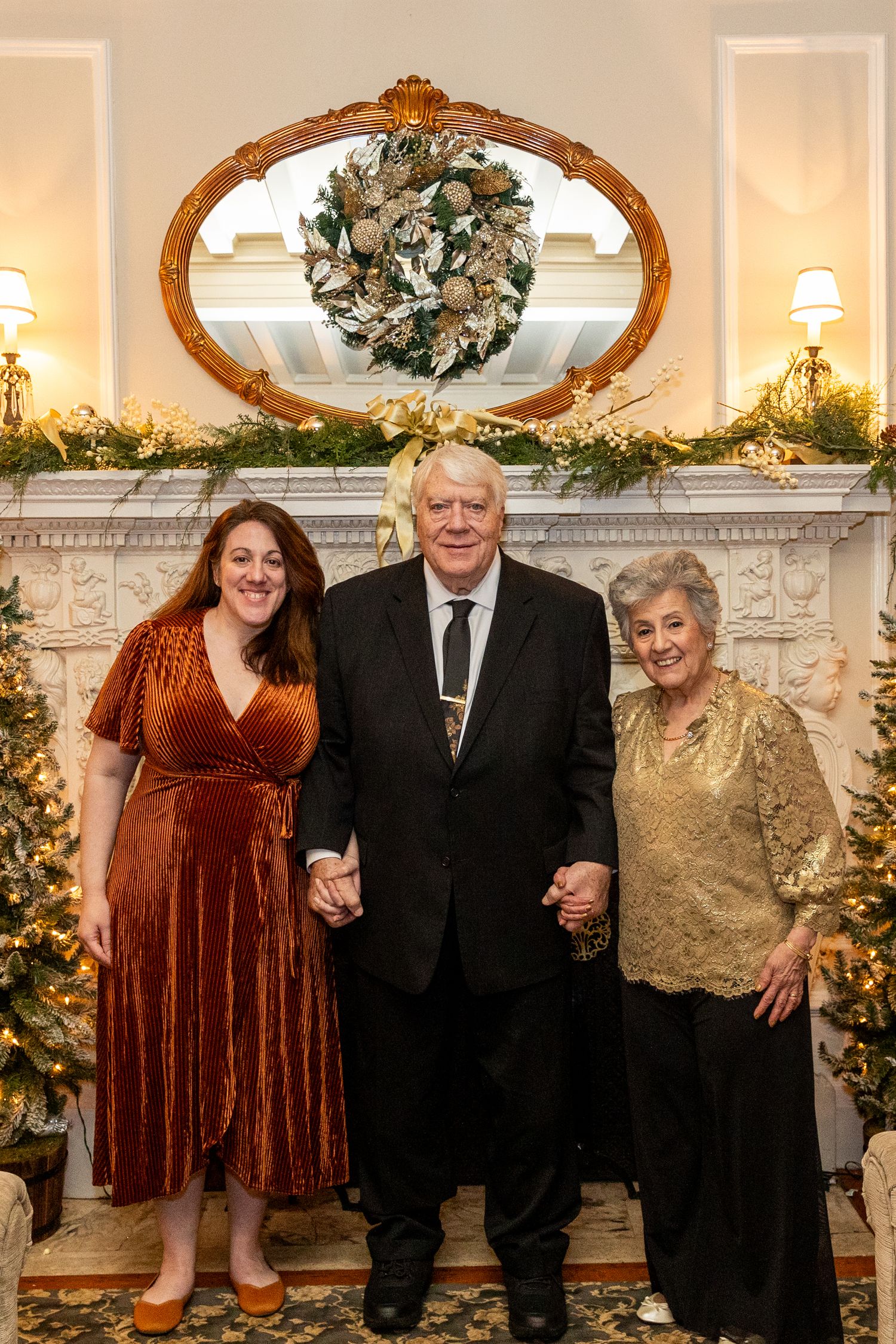 Three guests posing by decorated fireplace with Christmas wreath and trees at holiday celebration.