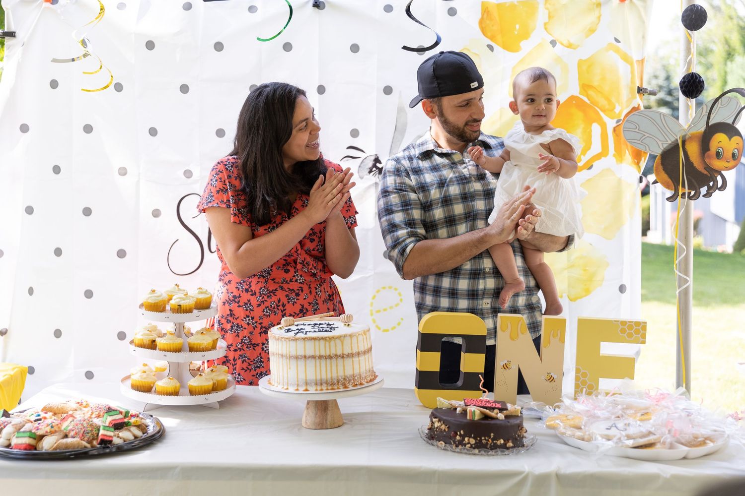 Family celebrating baby's first birthday with bee theme decorations, cake, and dessert table.