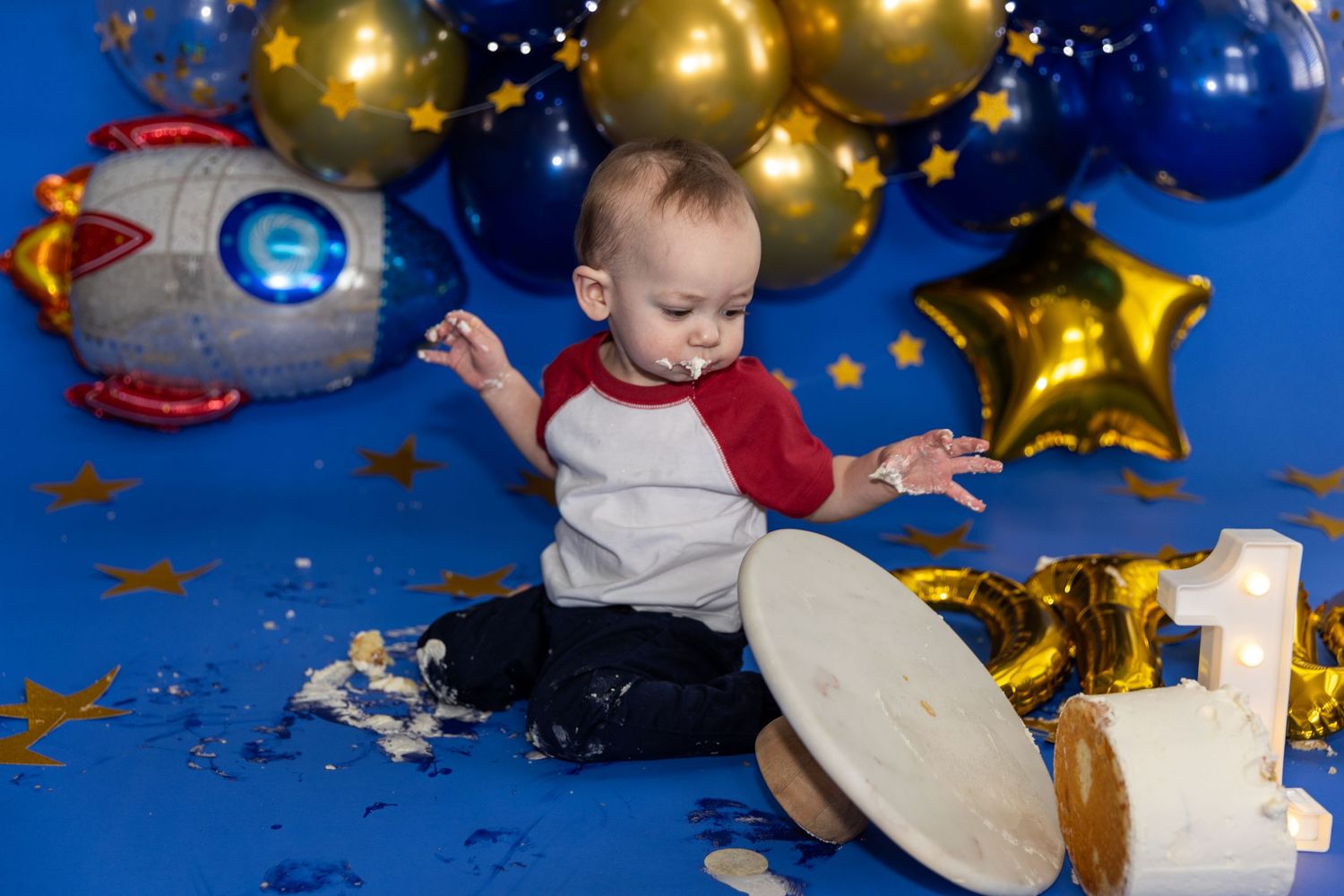 Baby boy smashing space themed first birthday cake with blue gold balloon backdrop and stars.