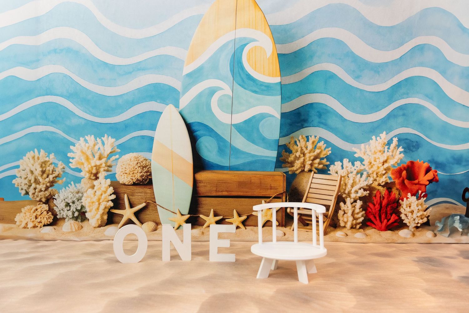 Beach-themed first birthday setup with surfboards, coral, starfish, and ONE letters on sandy floor.