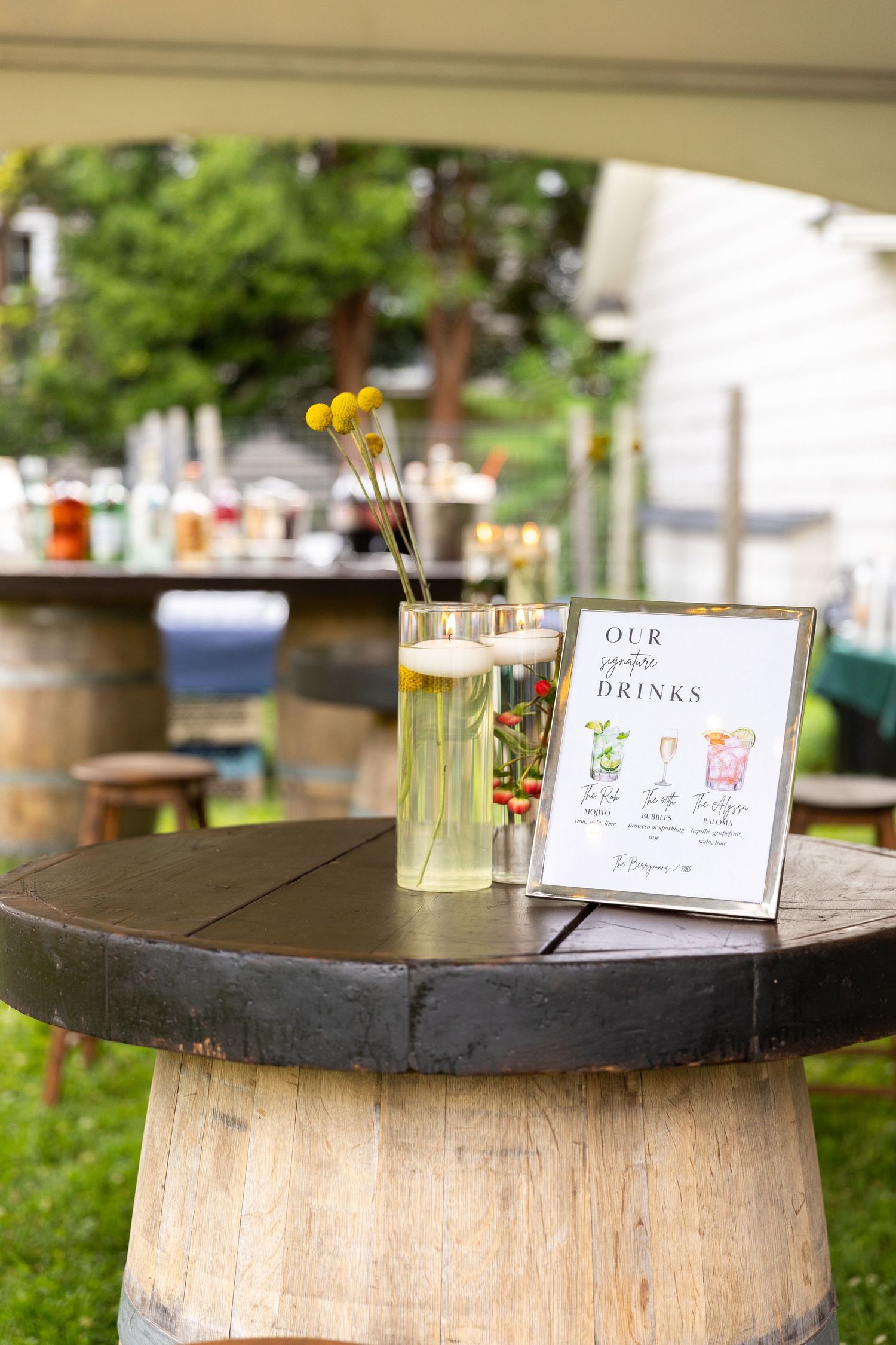 Rustic barrel cocktail table with signature drinks menu sign at an outdoor wedding reception.