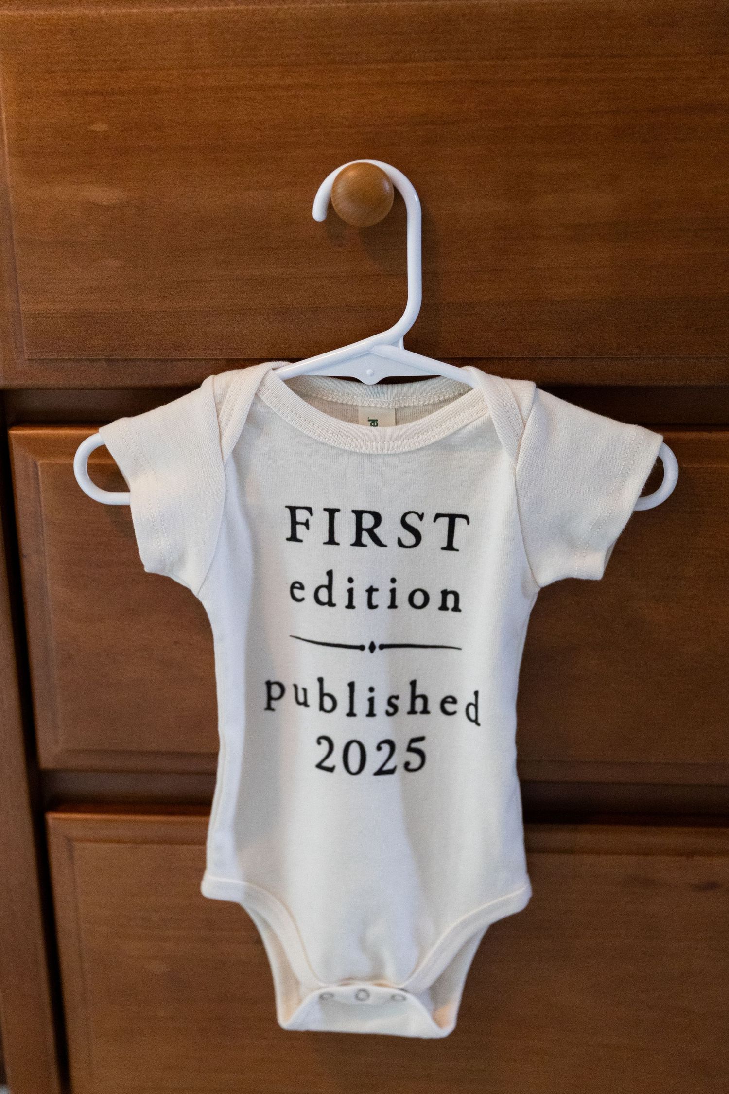 White baby onesie on hanger reading First Edition Published 2025 hanging on wooden dresser.