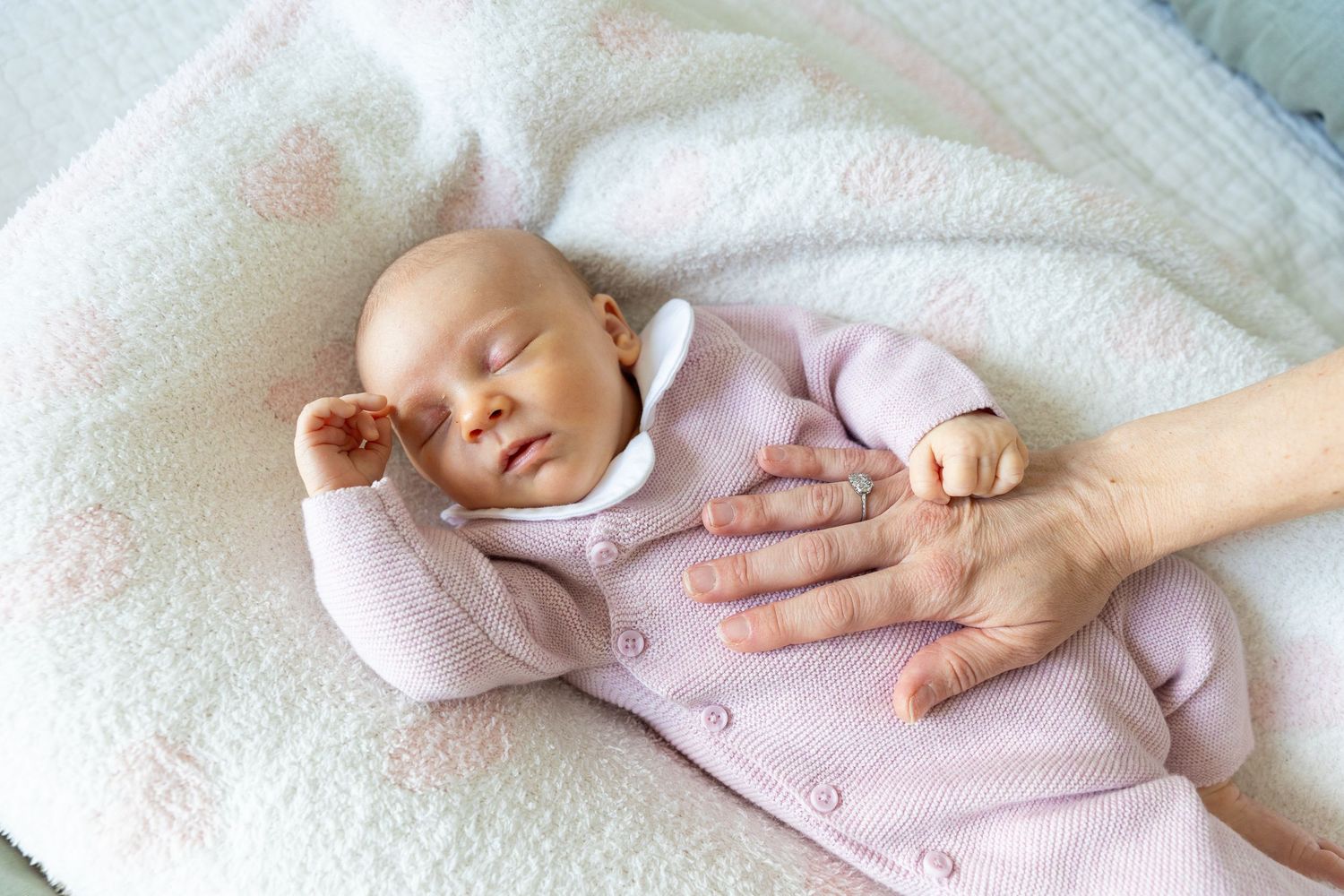 Sleeping newborn baby in pink outfit resting peacefully while parent's hand rests nearby.