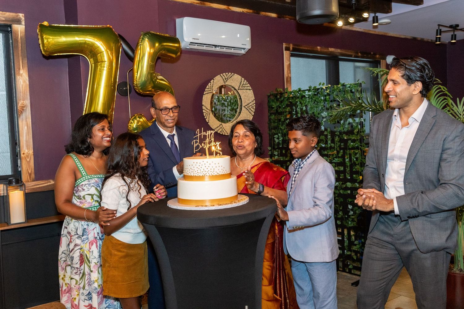 Family gathered around a 75th birthday cake with gold balloons at an indoor birthday celebration.