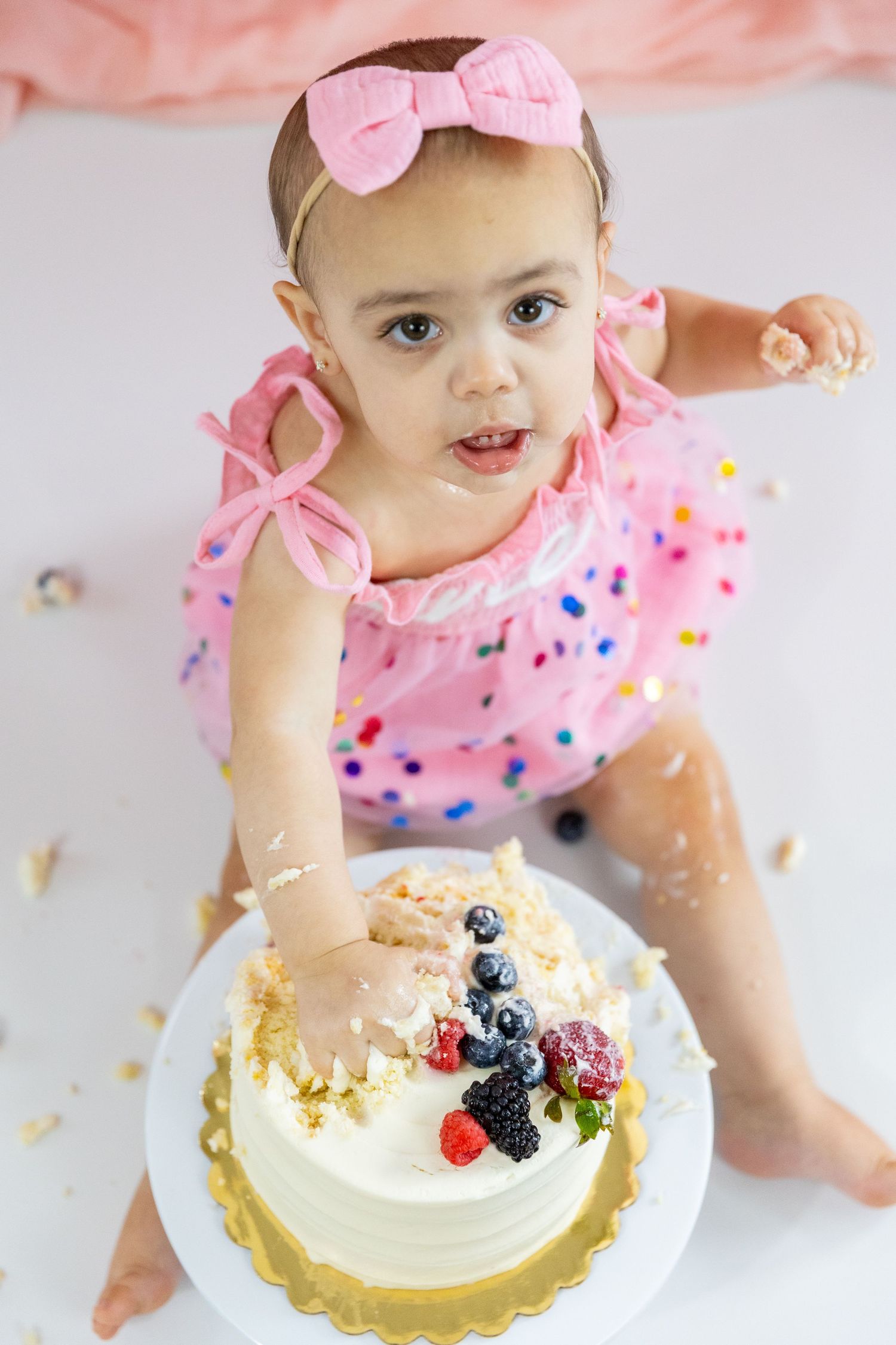 Adorable baby girl in pink polka dot outfit reaching into white berry-topped smash cake from above.
