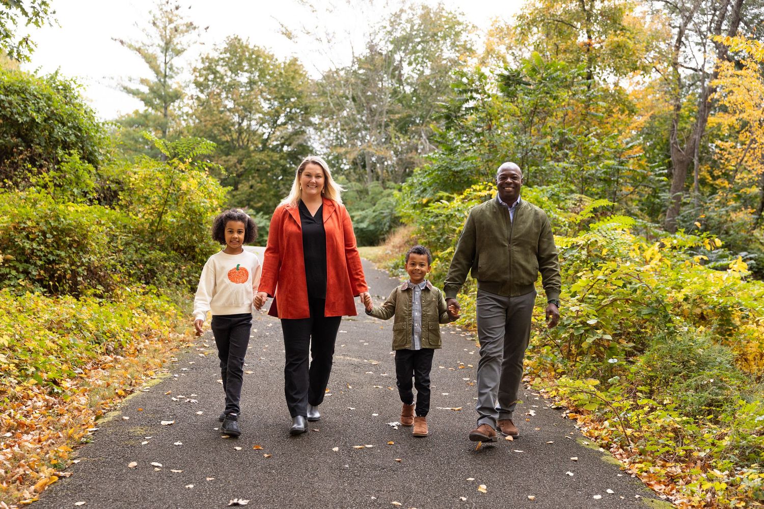 Happy interracial family of four walking hand in hand on tree-lined path during autumn foliage season.