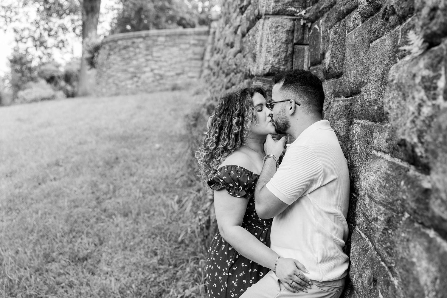 Black and white romantic couple photo leaning against stone wall outdoors in lush garden setting.