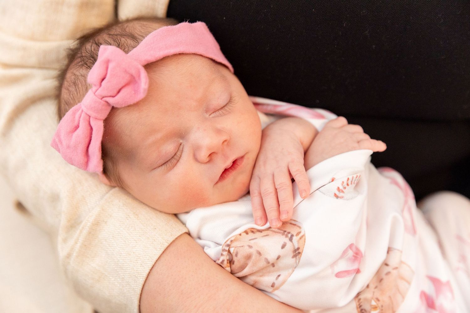 Newborn baby girl with pink bow headband sleeps peacefully while being cradled in loving arms.