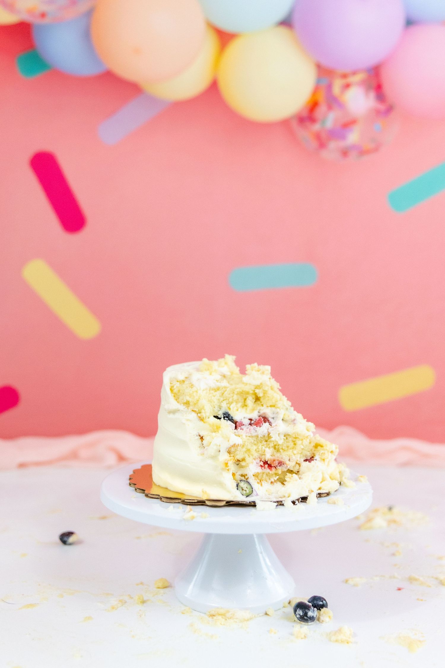 Smashed white birthday cake on pedestal stand with colorful balloon arch and confetti backdrop decoration.