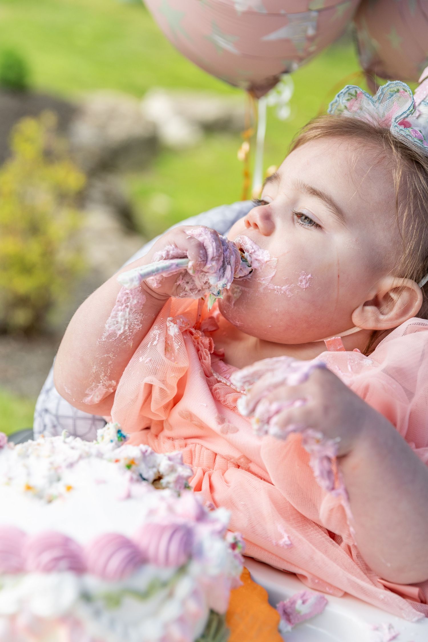 Adorable baby girl with frosting on her face enjoying her smash cake at an outdoor birthday party.
