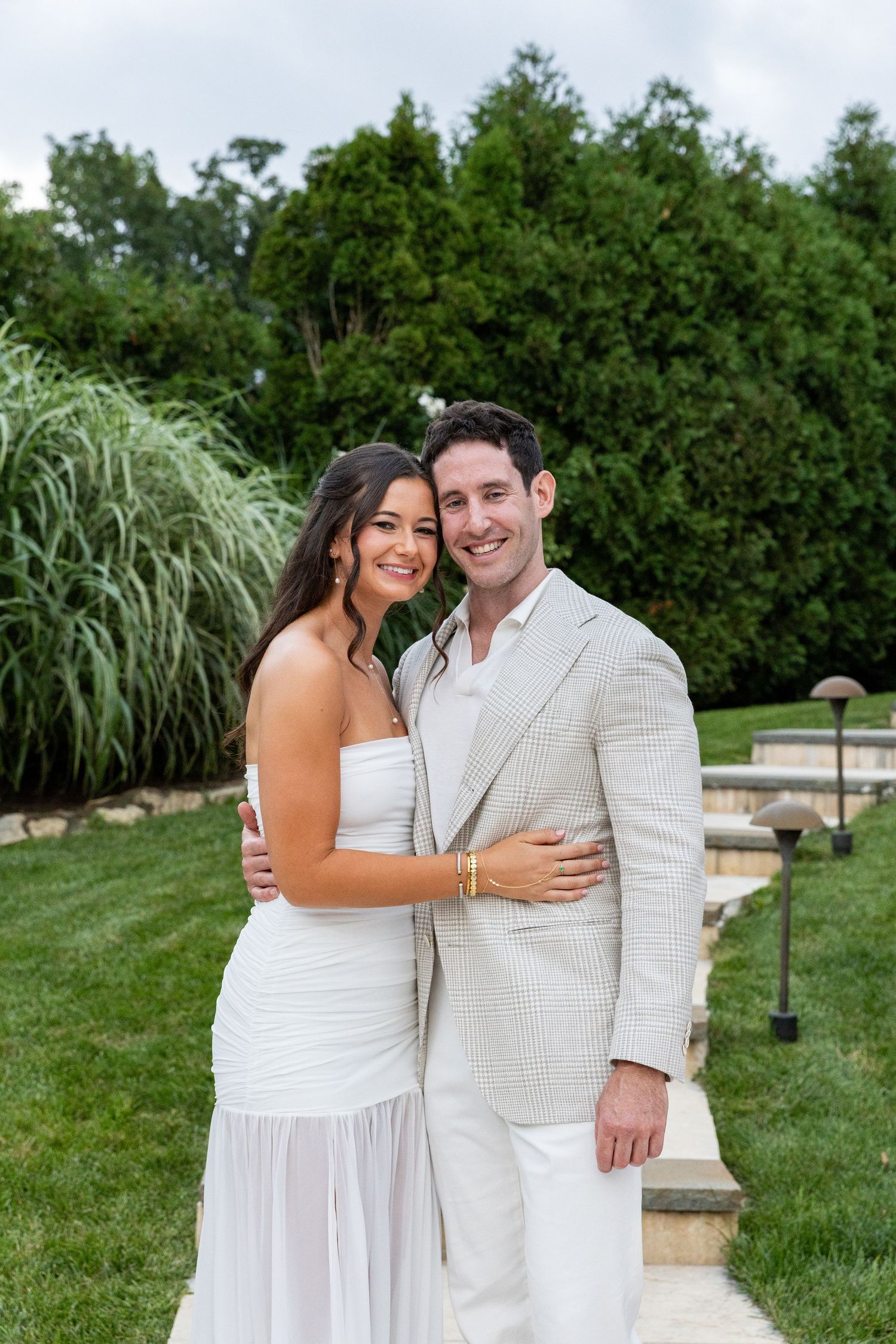 Smiling engaged couple posing together in white outfits in lush green garden setting.